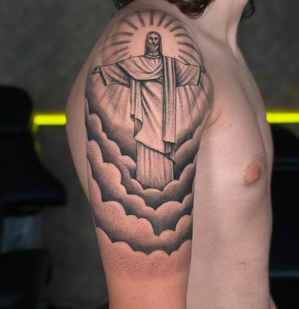 Christ Rédempteur dans les nuages, rayons de lumière, en réalisme noir et gris sur l’épaule, par Kako Tattoo à Genève
