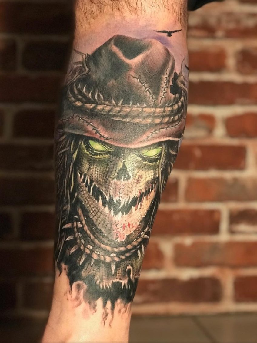 Épouvantail aux yeux verts sous un chapeau rapiécé, en réalisme couleur sur le mollet, par Kako Tattoo à Genève